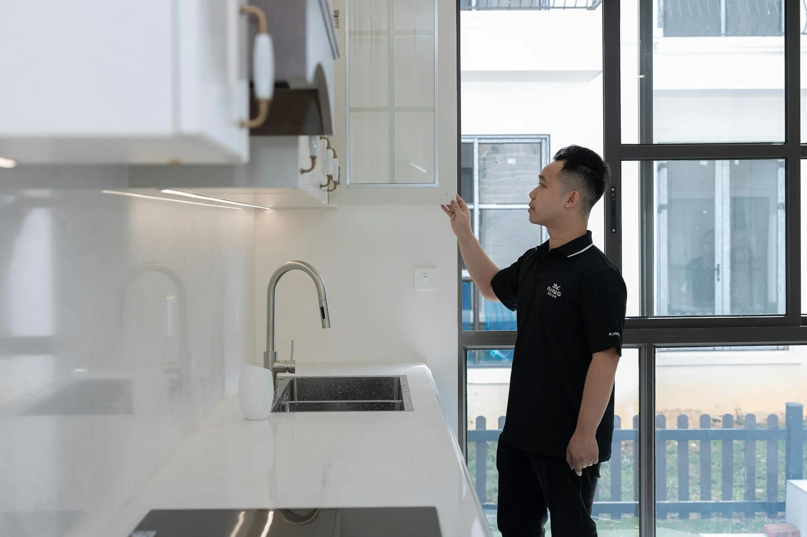 Người đàn ông mặc đồ đen đang kiểm tra tủ bếp trong căn hộ hiện đại có cửa kính lớn giúp phòng bếp thêm sáng. | Thiết kế nội thất 24
