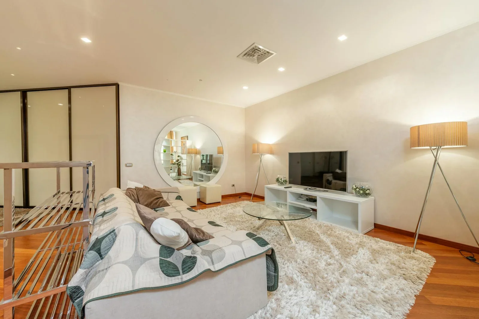 Sofa êm ái cùng bàn trà kính tròn trên thảm mềm gần tivi trong phòng khách rộng rãi đón ánh sáng dịu nhẹ. | Thiết kế nội thất 24